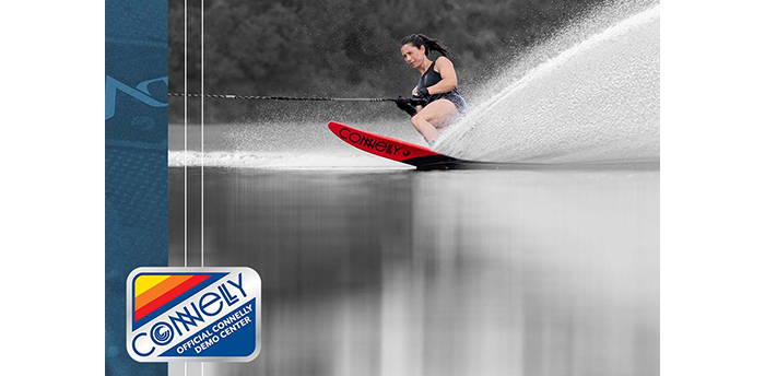 A female waterskier carves across the water on a red Connelly ski, creating a powerful spray as she leans into the turn.