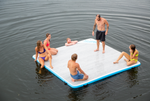 Side view of the Party Cove Dock Deluxe with people sitting and standing on its light woodgrain surface and blue border.