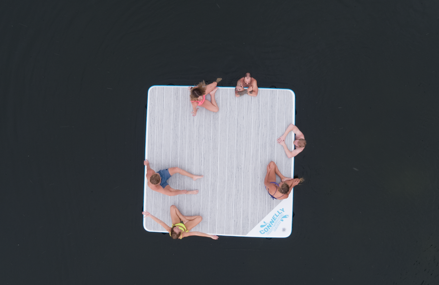 Overhead view of the Party Cove Dock Deluxe floating on water, featuring a light woodgrain surface and blue border.