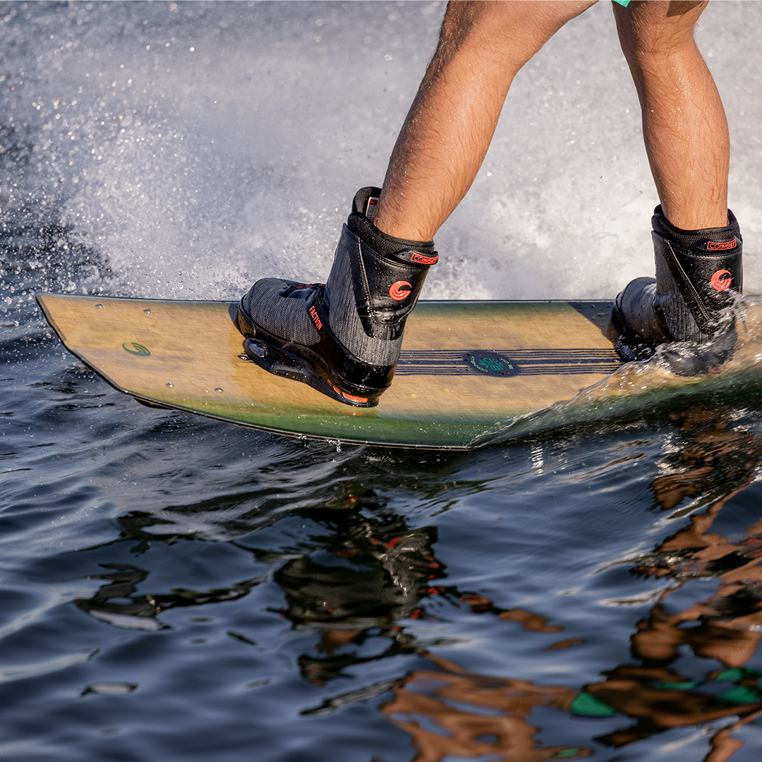 Close-up of a wakeboarder’s feet in bindings on the Bio wakeboard, skimming the water’s surface with a natural wood finish.