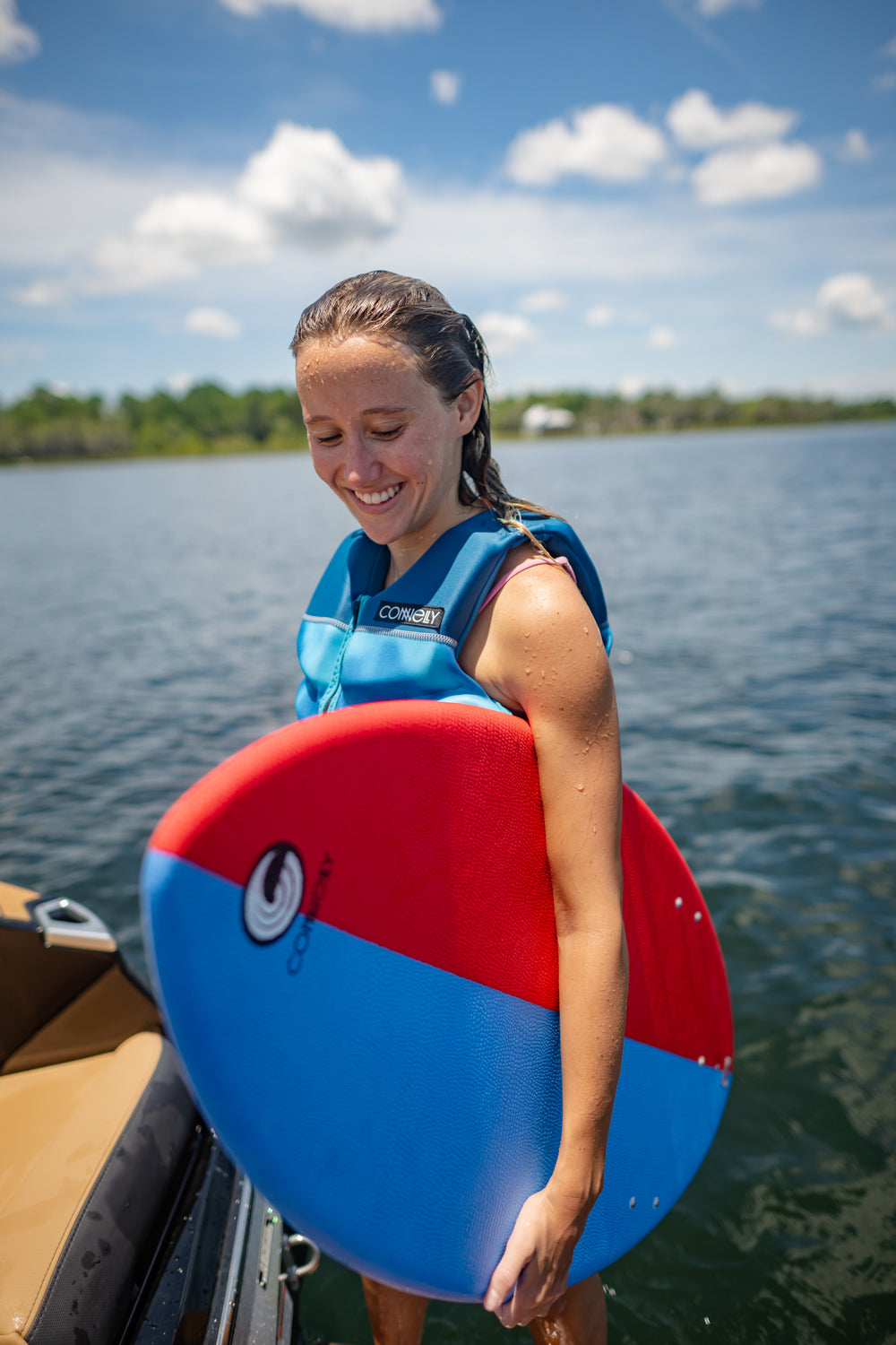 Young rider standing near the water holding the DASH Kid's Board, smiling while wearing a blue life vest.