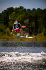 Wakeboarder mid-air on the Lotus wakeboard, wearing a black vest and pink shorts while gripping a tow rope over water.