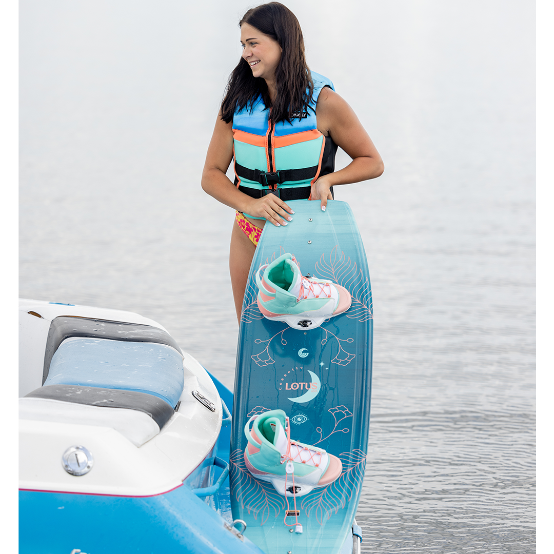 A young woman holding the Connelly Lotus wakeboard with attached pastel bindings near a boat on the water.