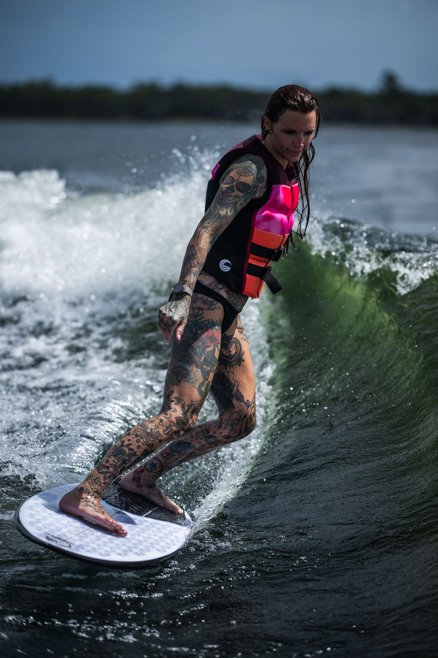 A female rider with tattoos and a pink life vest surfs a wave on the Ono wakesurf board.