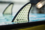 The Ono wakesurf board’s fin with a honeycomb design is partially submerged in water.