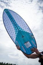 A person holds the Ono wakesurf board, showcasing its blue and white geometric traction pad and fin slots.