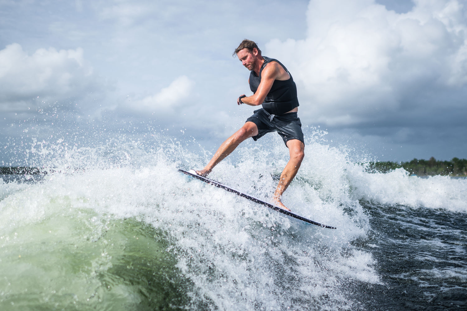 A male rider carves through a wave on the Ono wakesurf board, wearing a black vest and gray shorts.