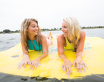 Close-up view of two women lying on the yellow Party Cove Island float, partially submerged in water and smiling.