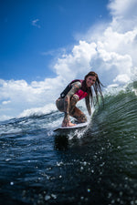 A female surfer in a pink life vest carves a wave on the Seer wakesurf board, crouching with a smile.