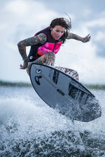 A female surfer launches into the air on the Seer wakesurf board, showcasing its sleek gray base with black accents.