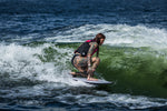 A female surfer crouches low while riding the Seer wakesurf board, maintaining balance on a green wave.