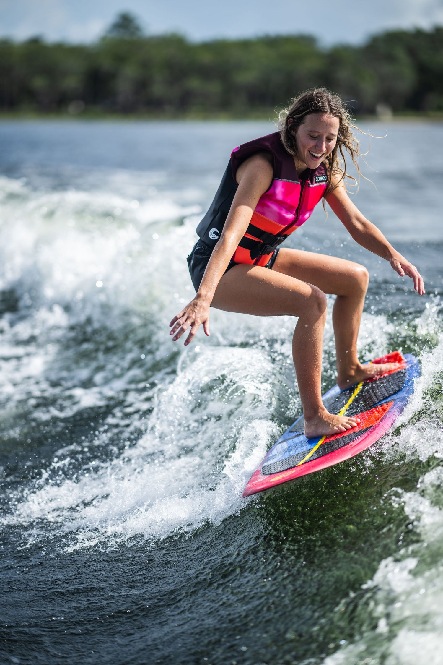 Woman in a pink and black life vest wakesurfing on the SPARK board, smiling with arms out for balance.