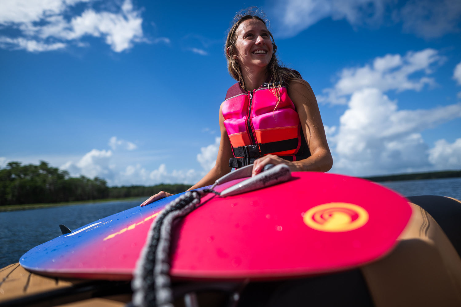 Woman in a pink and black life vest holding the SPARK wakesurf board on a boat with a lake in the background.