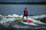 Rear view of a woman wakesurfing on the SPARK board, creating a foamy white wave trail on the lake.