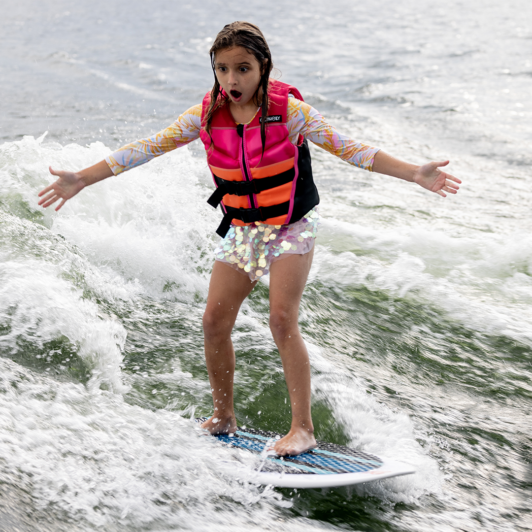 Young girl in a pink life vest rides the Scout wakesurf board, arms outstretched while balancing on a wave.