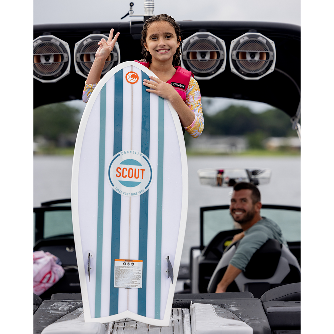 Young girl stands on a boat platform, smiling while holding the Scout wakesurf board vertically.