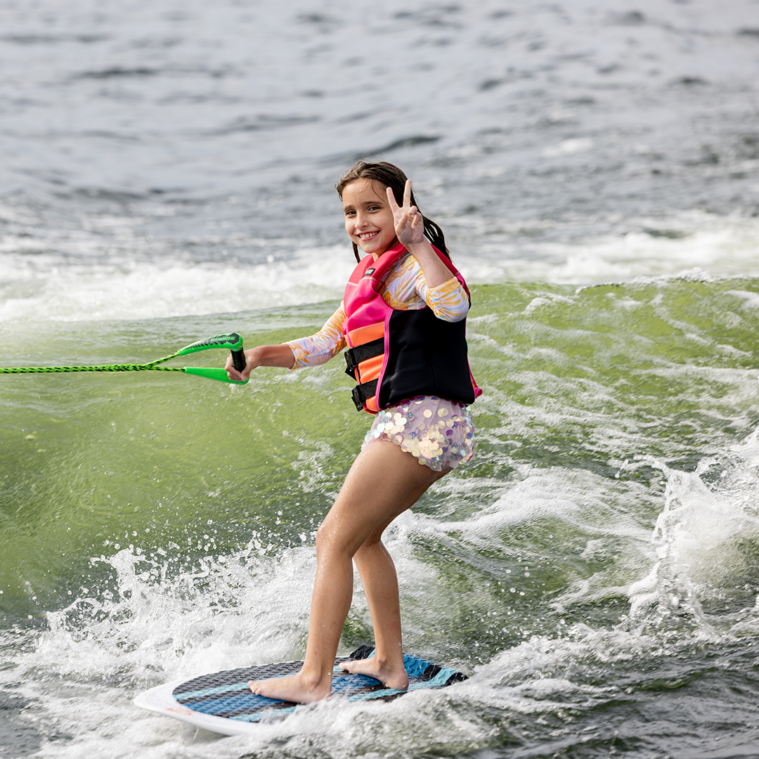 Young girl smiles and holds up a peace sign while riding the Scout wakesurf board with a tow rope.