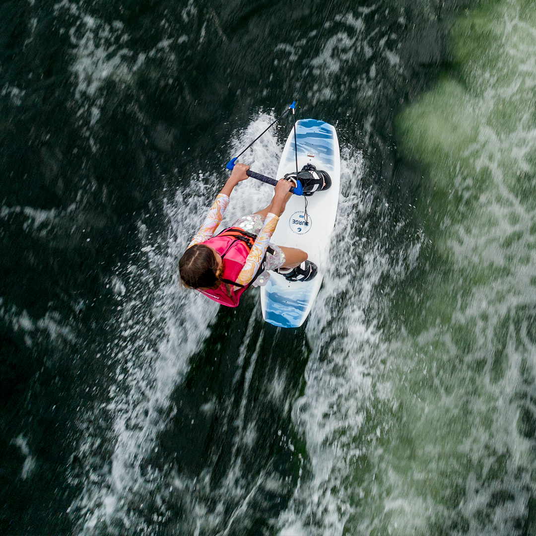 A young rider in a pink vest wakeboarding on the Surge, gripping a handle while carving through the water.