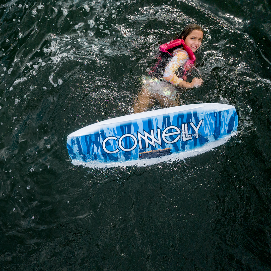 A young rider in a pink vest floating in the water next to the Surge wakeboard.