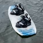 Surge wakeboard floating on the water with black and white bindings attached.