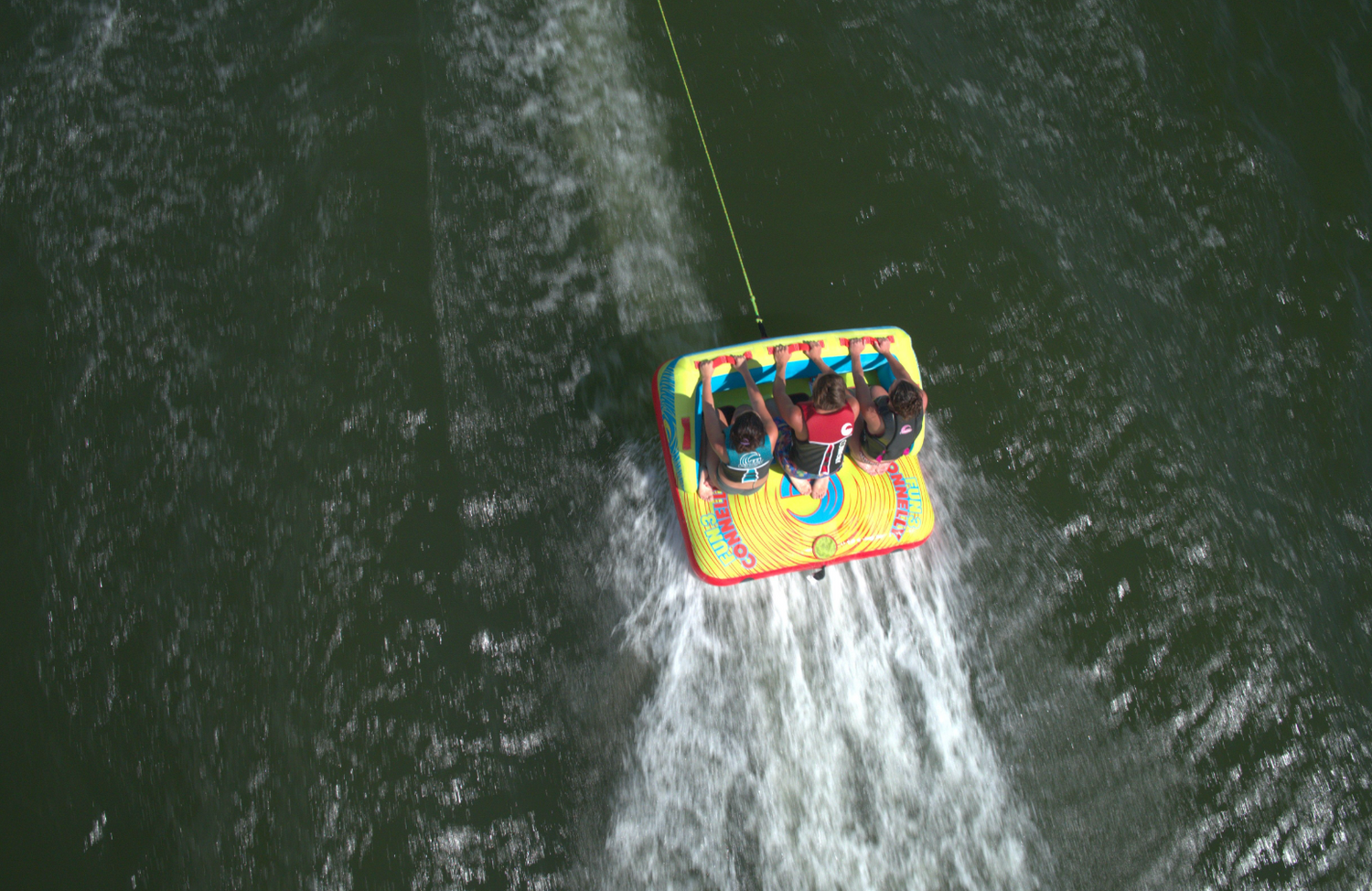 Fun 3 towable tube with three riders gripping handles as they are pulled across the water, leaving a wake behind them.