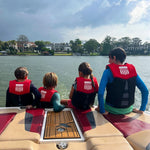 Four children sitting on a boat deck wearing Junior MC Neo Vests, facing the water with houses visible in the background.