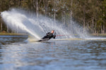 A waterskier carves through the water on a Connelly 2023 DV8 ski, creating a spray against a backdrop of trees and water.