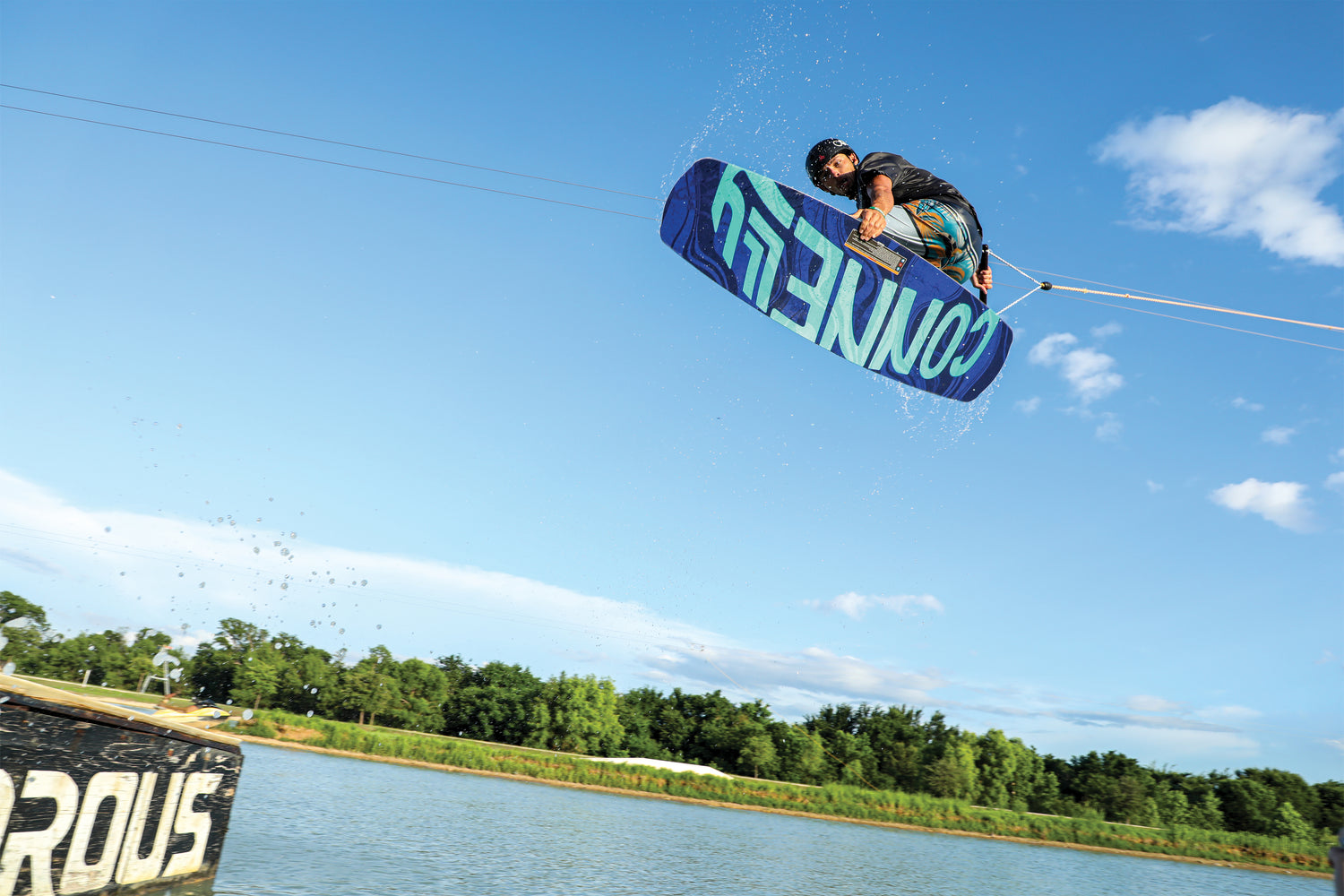 The 2024 Groove wakeboard in mid-air, showcasing its bold blue and teal graphics with dynamic water spray.