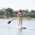 Woman paddleboarding on a calm lake, wearing a blue top and pink bottoms, balancing on a Classic SUP.