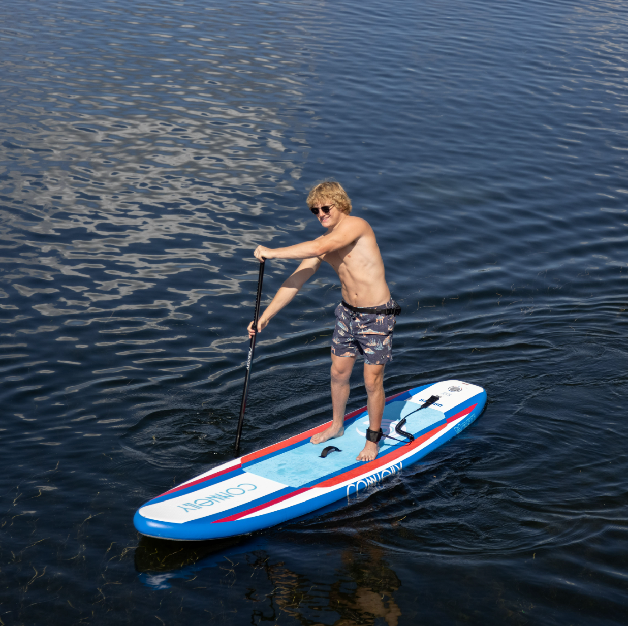 A man in floral shorts paddles on the Drifter iSUP, maintaining balance while navigating the water.