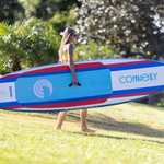 A woman in a yellow swimsuit carries the Drifter iSUP with a center handle, walking barefoot on grass.